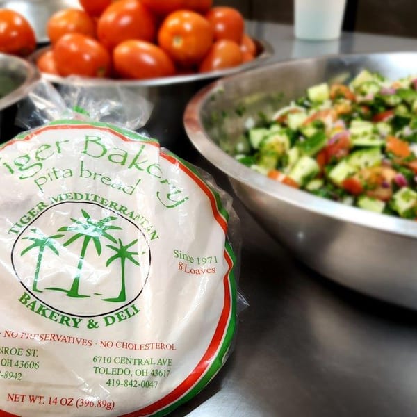 Tiger Bakery pita bread package next to bowls of fresh cucumbers, tomatoes, and salad.