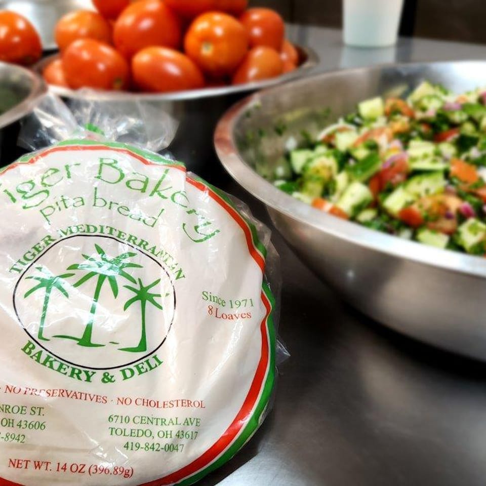 Tiger Bakery pita bread package next to bowls of fresh cucumbers, tomatoes, and salad.